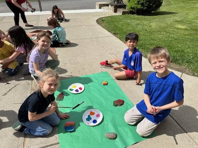 students on sidewalk painting rocks on a green piece of paper.