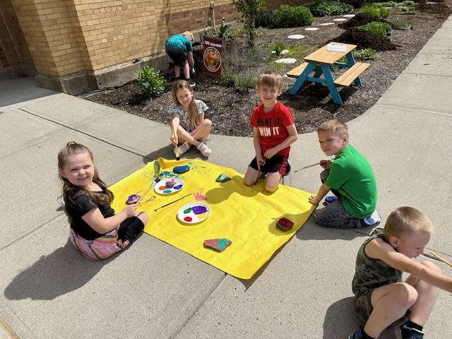 students on sidewalk painting rocks on a yellow piece of paper.