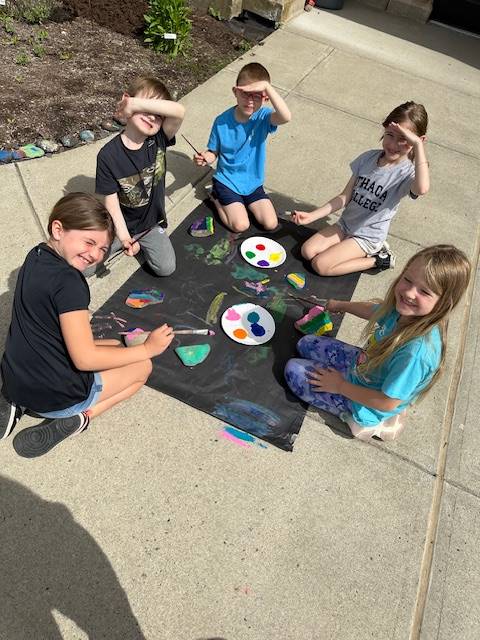 students on sidewalk painting rocks on a black piece of paper.