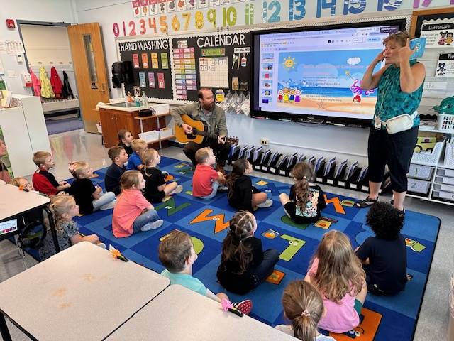 1 adult on guitar, 1 adult standing and students watching on rug