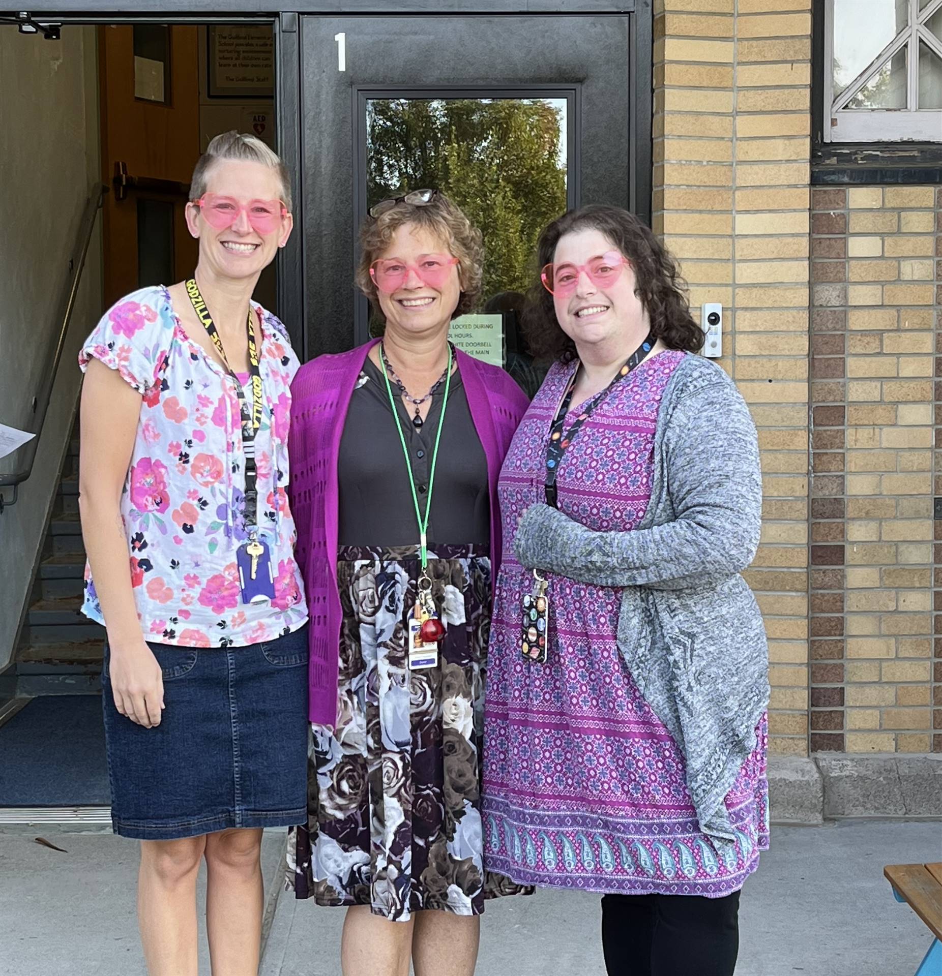 3 adults with pink heart shaped sunglasses in front of school door