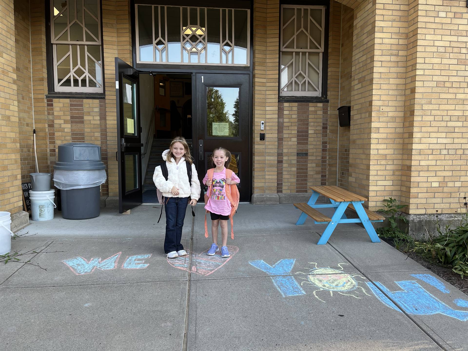 2 students standing on a sidewalk chalk heart in front of a school doorway