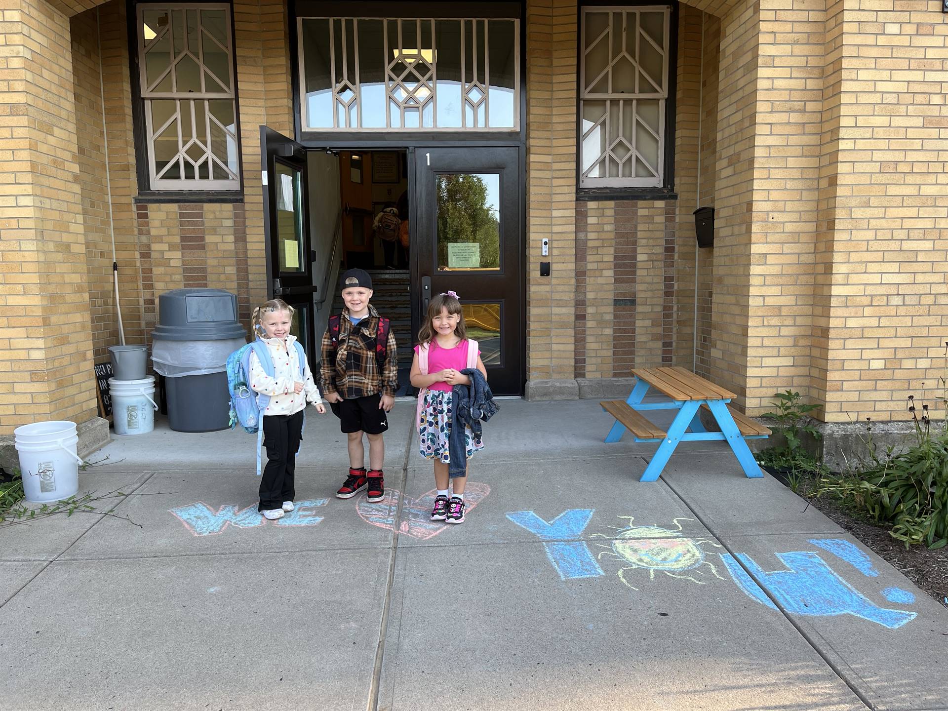 3 students standing on a sidewalk chalk heart in front of a school doorway