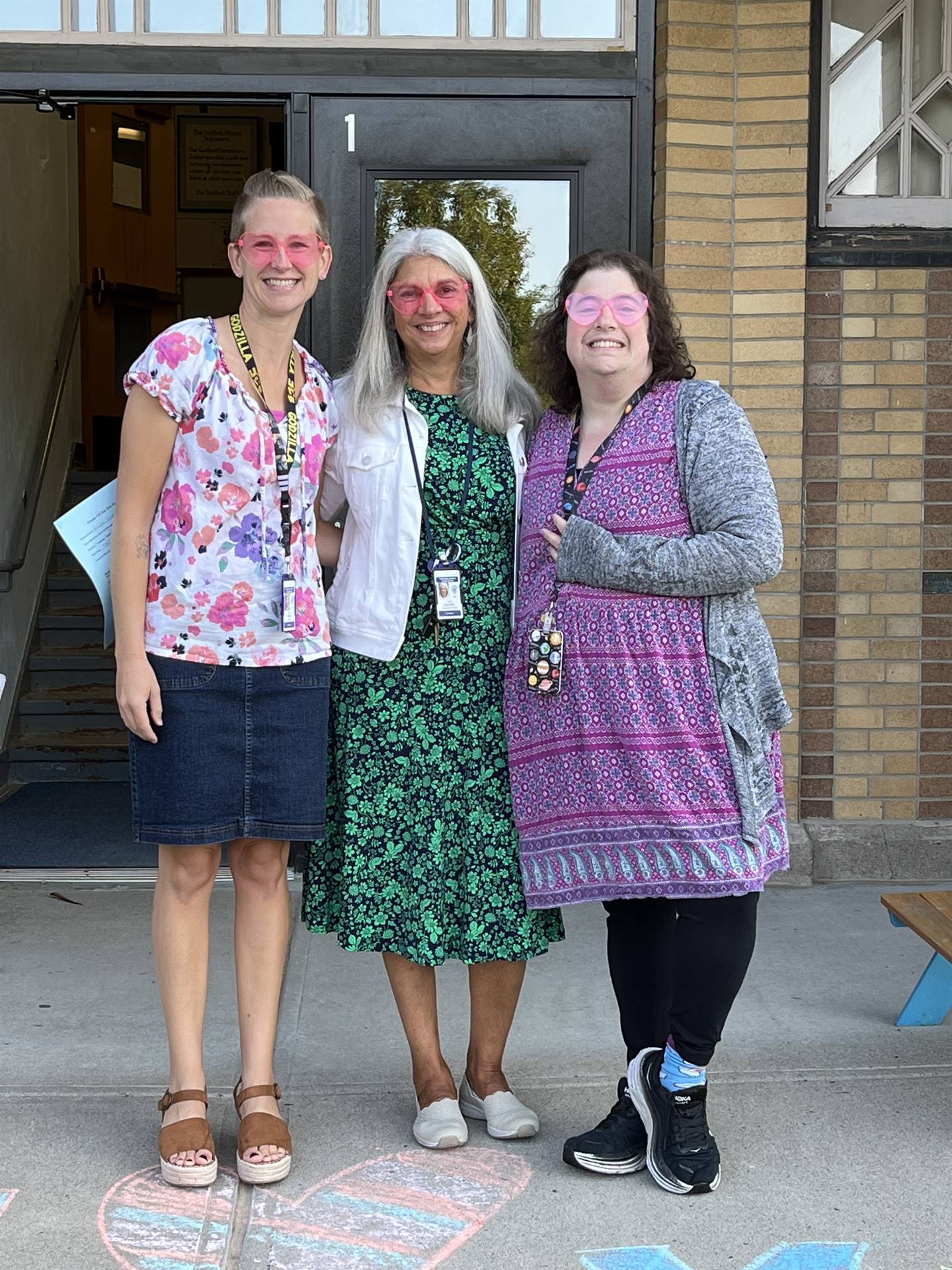 3 adults with pink heart shaped sunglasses in front of school door